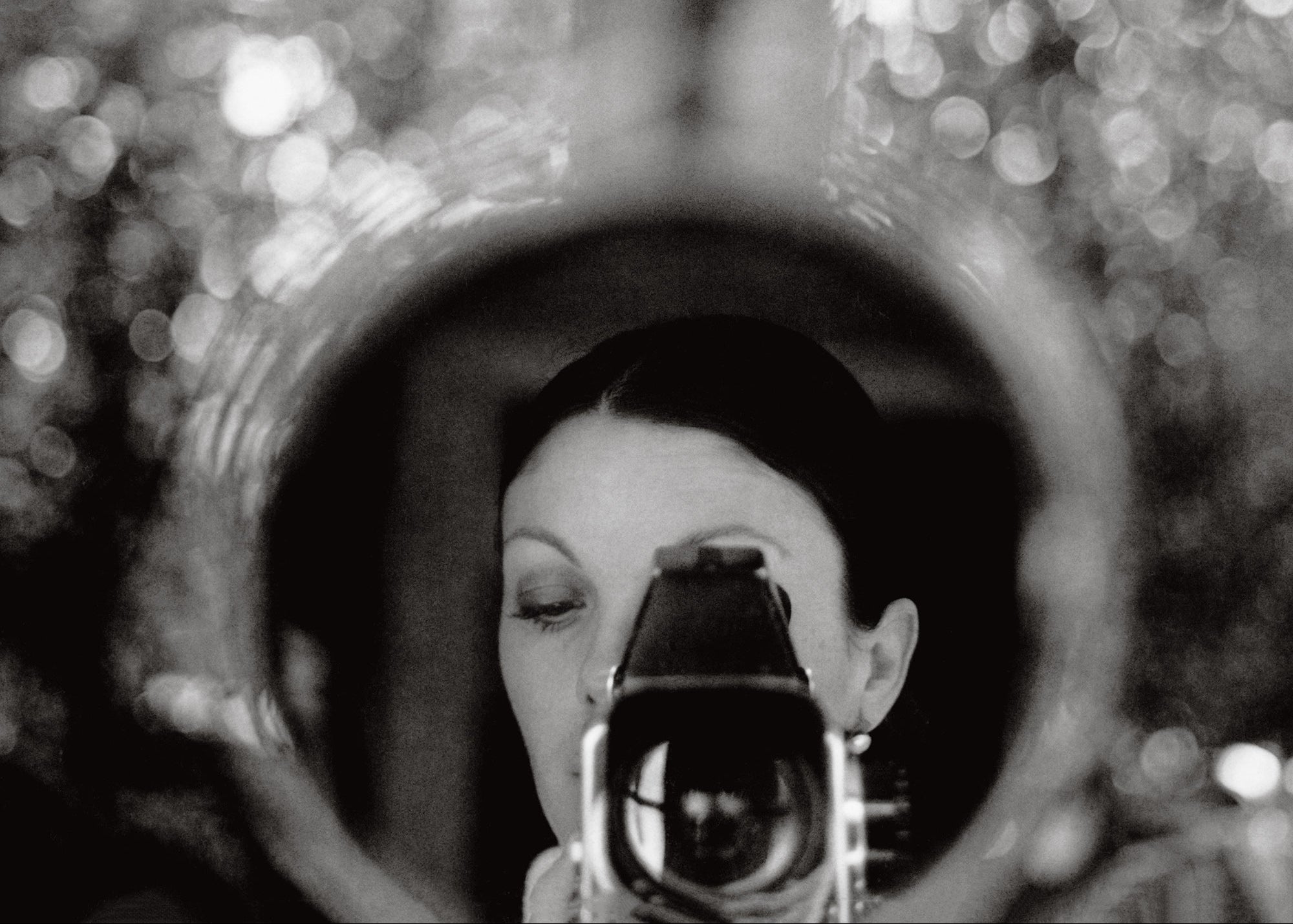 Black and white photograph of a woman holding a vintage camera in front of her face, framed by a circular reflective surface with a blurred bokeh background.