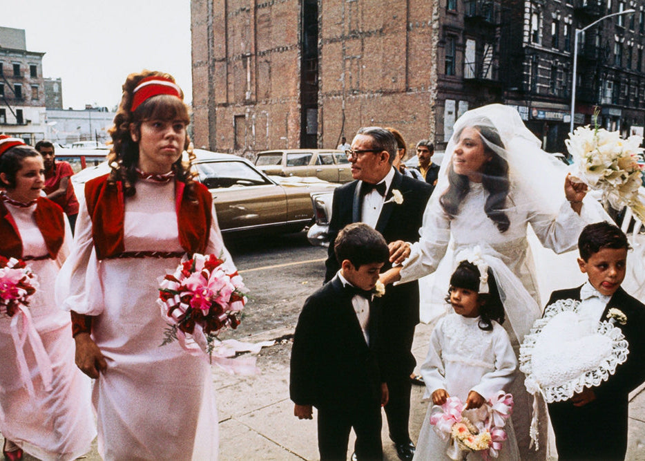 A candid street photograph of a wedding party walking down a city sidewalk. The bride in a white wedding gown and veil holds a bouquet, surrounded by bridesmaids in white dresses with red shawls, a ring bearer in a white gown, and two young flower boys in black suits.