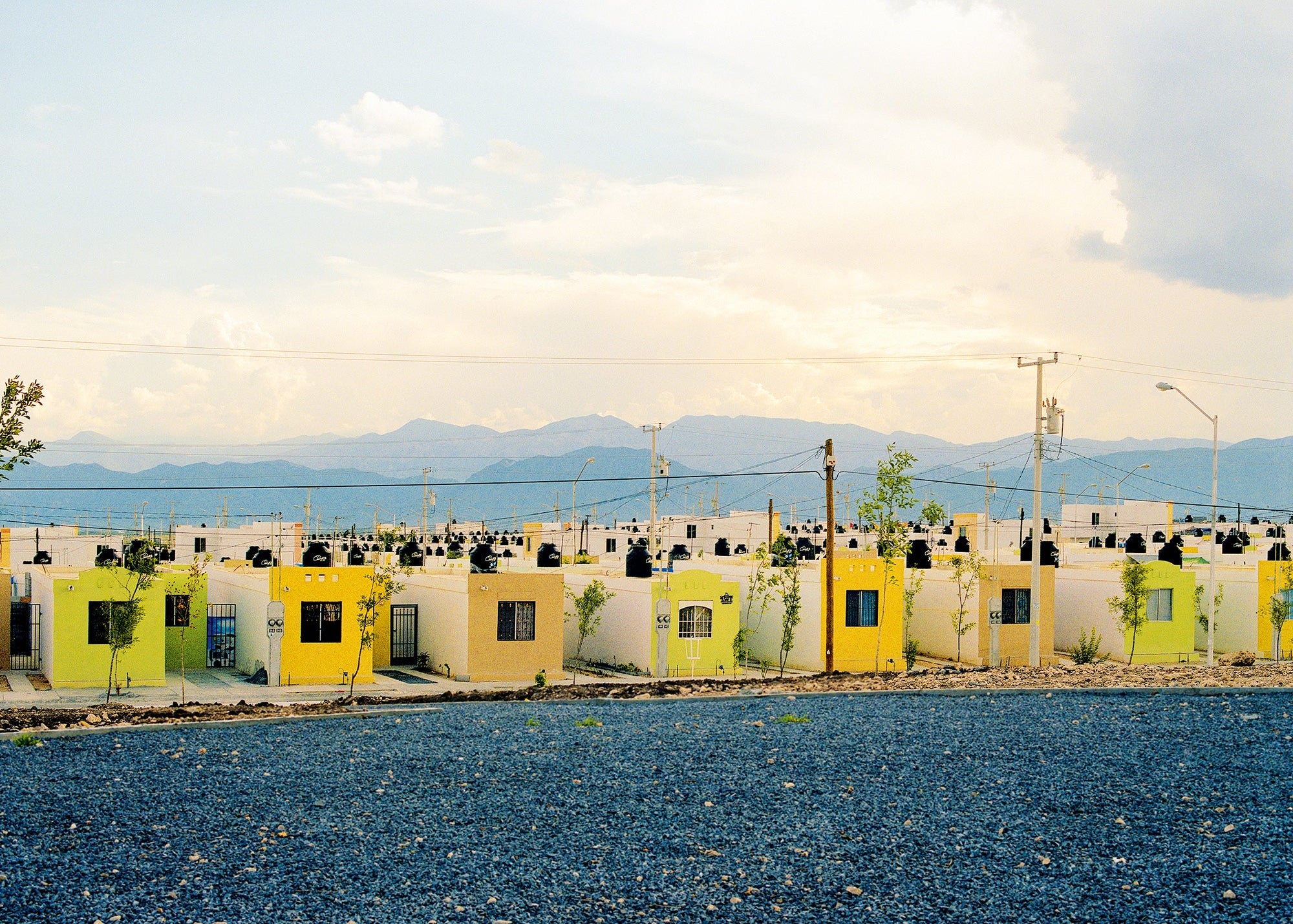 A row of small white and yellow buildings with dark roofs sits behind a gravel foreground, with mountains and a cloudy sky in the background.